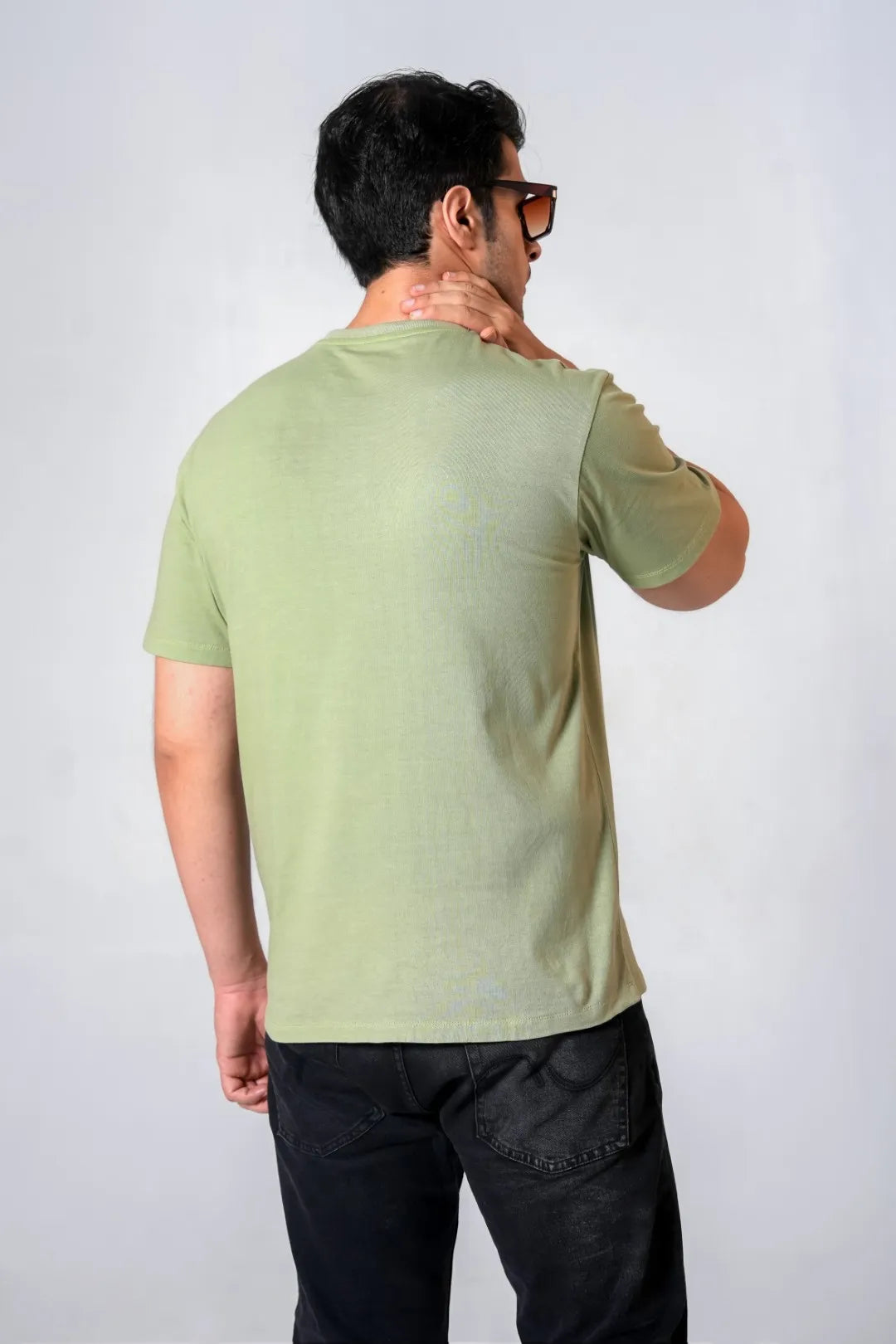 Back view of man in olive green cotton t-shirt and black jeans, one hand on neck, standing in a clean, modern studio environment