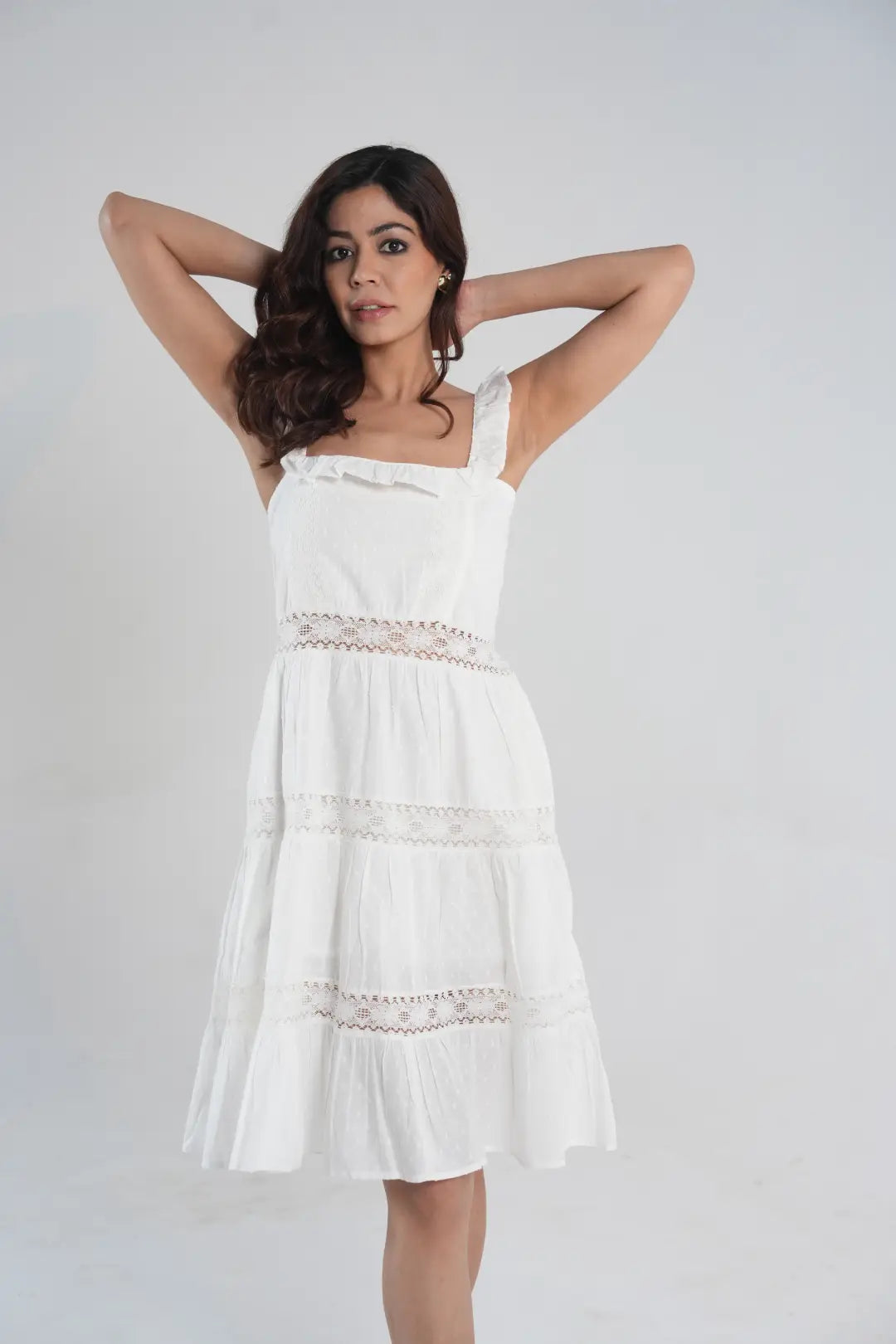 Posing against a light gray background, a woman with wavy brown hair models a white cottagecore-style sundress complete with a tiered skirt, square neckline, ruffled straps, and delicate lace detailing.