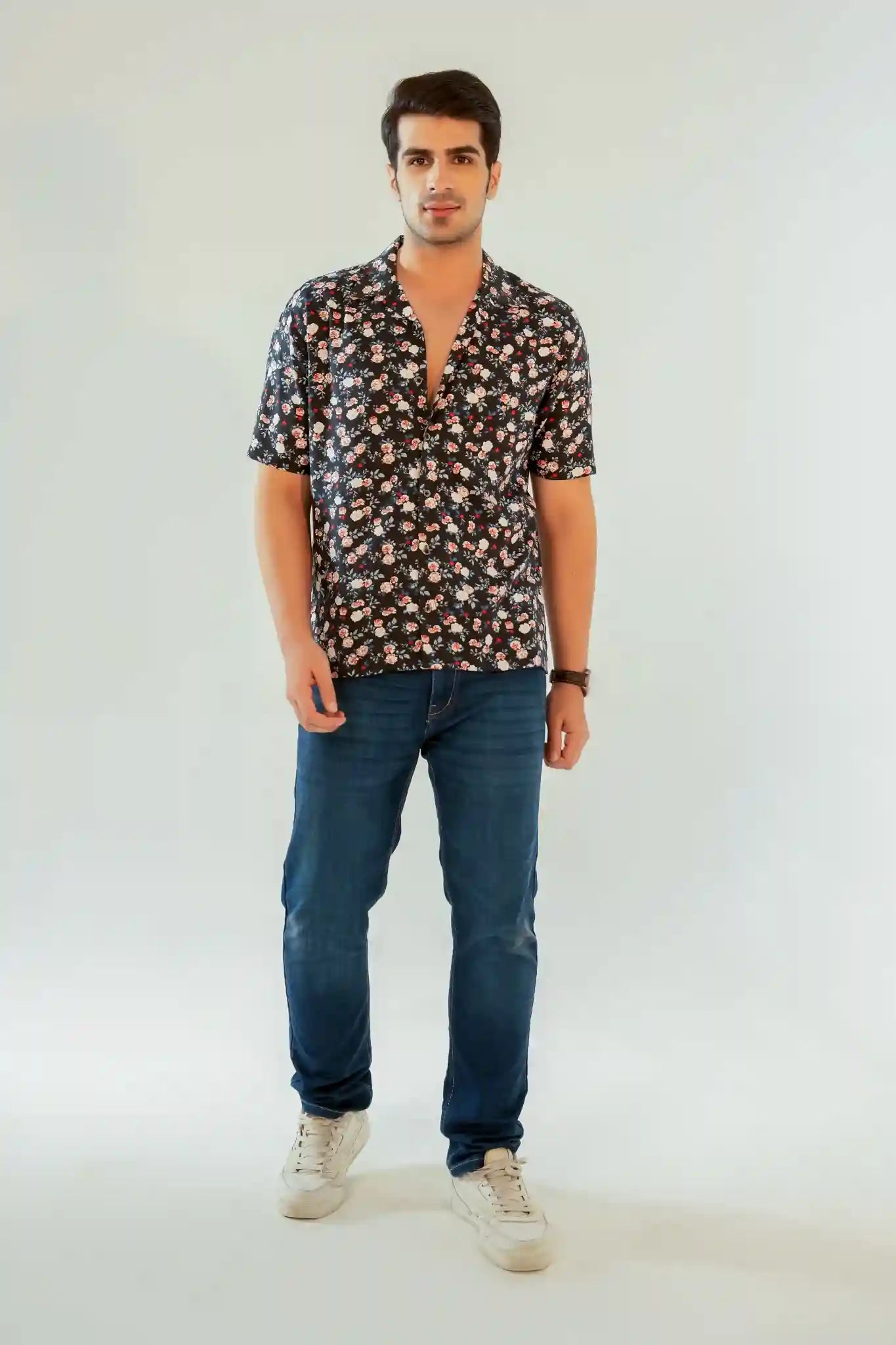Man standing in a navy floral Cuban collar shirt with short sleeves, styled with blue jeans and white sneakers on a plain backdrop