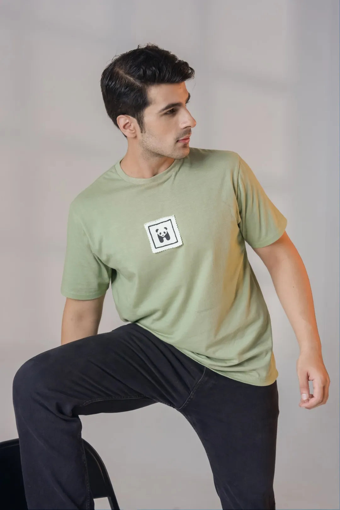 Man posing in an olive green cotton t-shirt with embroidered panda patch on chest, paired with black jeans, styled in a relaxed look