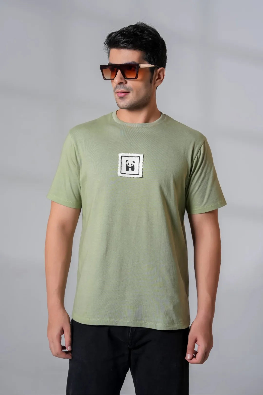 Man in olive green t-shirt with panda patch, accessorized with oversized brown sunglasses and black jeans for a casual street look