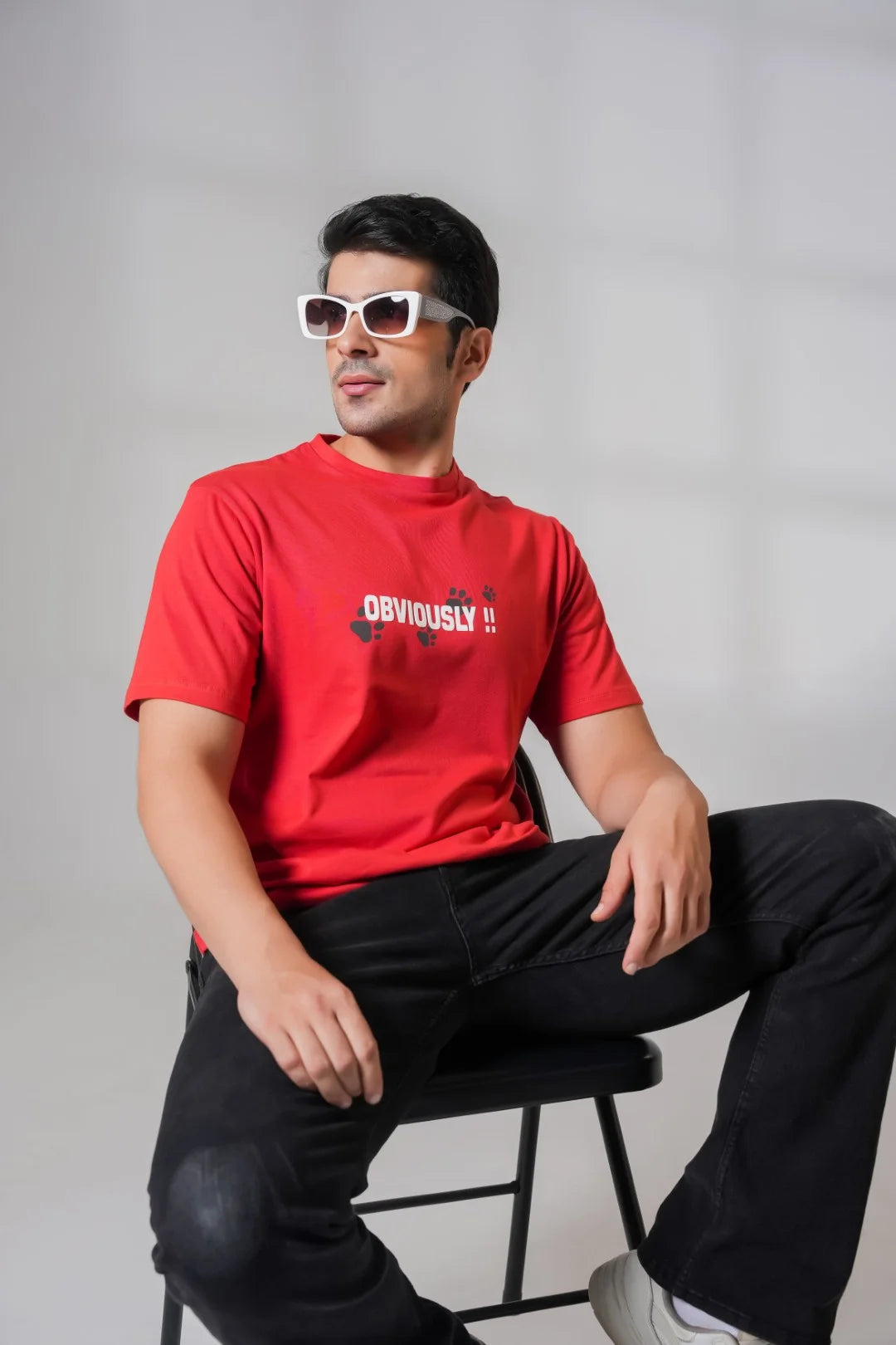 Man sitting on a black chair wearing a red graphic t-shirt with white sunglasses, styled with black jeans for a cool casual look