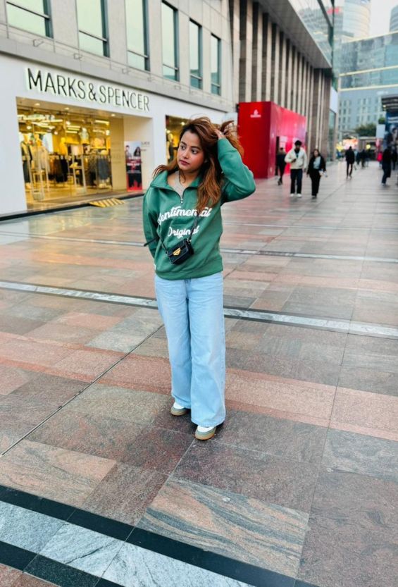 Woman in a green sweatshirt and light blue jeans standing in front of a Marks & Spencer store - Drapepanda